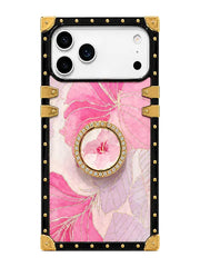 A protective phone case featuring a pink and lavender floral print, a central circular crystal-embellished grip ring, and a black border with gold-tone metal studs and corner protectors.