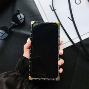 Hand holding a smartphone with a black and gold case on a dark surface with sunglasses and a magazine.