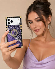 A woman takes a mirror selfie showing off a custom phone case. The case features a purple leopard print top section, a purple glitter lower section, a white rhinestone-embellished diagonal stripe, and black trim with gold studs and corner accents.
