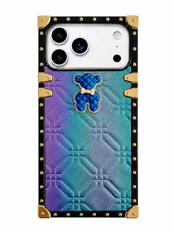 A protective phone case designed to look like a luxury trunk, featuring a black faux leather body with gold-tone studs, corner protectors, and a quilted pattern. The center has a smooth iridescent gradient transitioning from purple to teal, adorned with a blue quilted teddy bear charm with a gold bow.