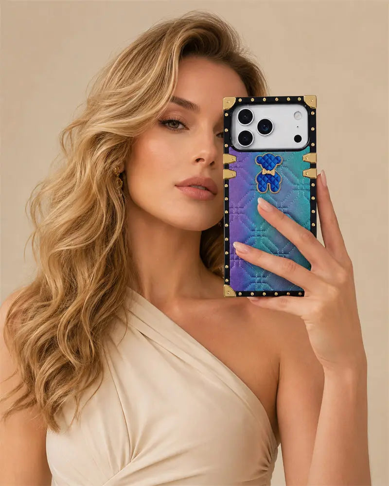 A woman in a beige one-shoulder dress holds up a smartphone case in a mirror selfie. The case features a gold and black studded border, a blue and purple holographic bear design, and a gold bear emblem in the center.