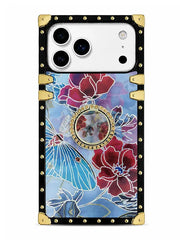 A luxury-style phone case featuring a black leather-look border with gold studs and corner protectors, a central circular mirror, and a vibrant floral and butterfly pattern in red, blue, and purple.