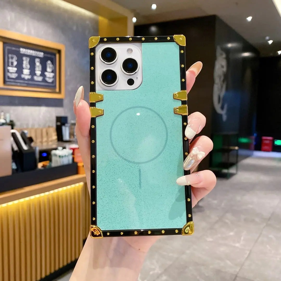 A hand holding a light blue magnetic phone case with a black and gold faux-leather border, shown in a cafe setting.