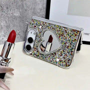 A silver glitter flip phone case with a heart-shaped mirror, styled on a white surface with a red lipstick in the foreground.