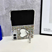 A silver flip-style iPhone Duo case with a glittery multicolor back, a heart-shaped mirror cutout, and a small dangling charm, displayed upright on a white surface against a gray wall.uo