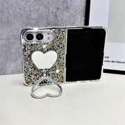 A silver foldable phone case covered in multicolor rhinestones, featuring a heart-shaped mirror on the back and a matching heart-shaped stand in front, displayed on a white surface against a gray textured background.
