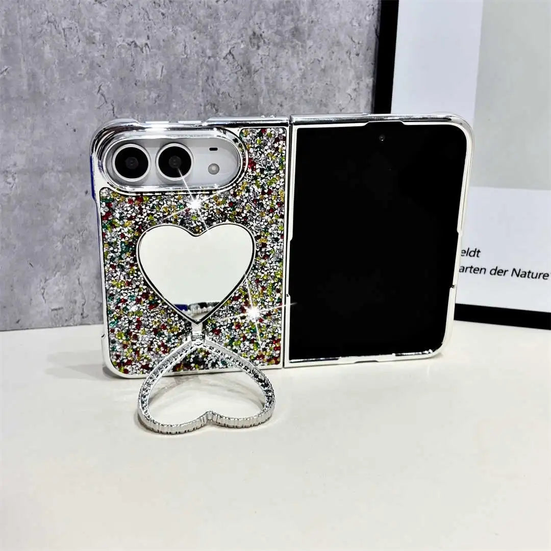 A silver foldable phone case covered in multicolor rhinestones, featuring a heart-shaped mirror on the back and a matching heart-shaped stand in front, displayed on a white surface against a gray textured background.