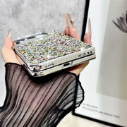 A hand with long nude manicured nails holds a slim rectangular phone case covered in dense multicolor rhinestones and framed by a shiny silver metallic border. The hand is wearing a sheer black long-sleeve top, with a gray textured wall and a framed print in the background.