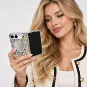 A woman holds a silver flip phone with a glittery multicolor case featuring a heart-shaped mirror. She is wearing a cream tweed jacket with black trim and gold buttons against a clean white background.