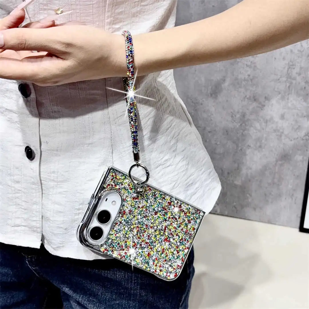 A close-up of a person wearing a sparkly phone case covered in multicolor glitter, with a matching glitter wrist strap attached. The person is wearing a white button-up shirt and dark blue jeans, standing against a gray textured wall.
