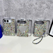 Three silver phone cases covered in multicolor rhinestones are displayed on a white surface against a gray concrete wall. One case features a heart-shaped mirror, and another has a matching rhinestone wrist strap.