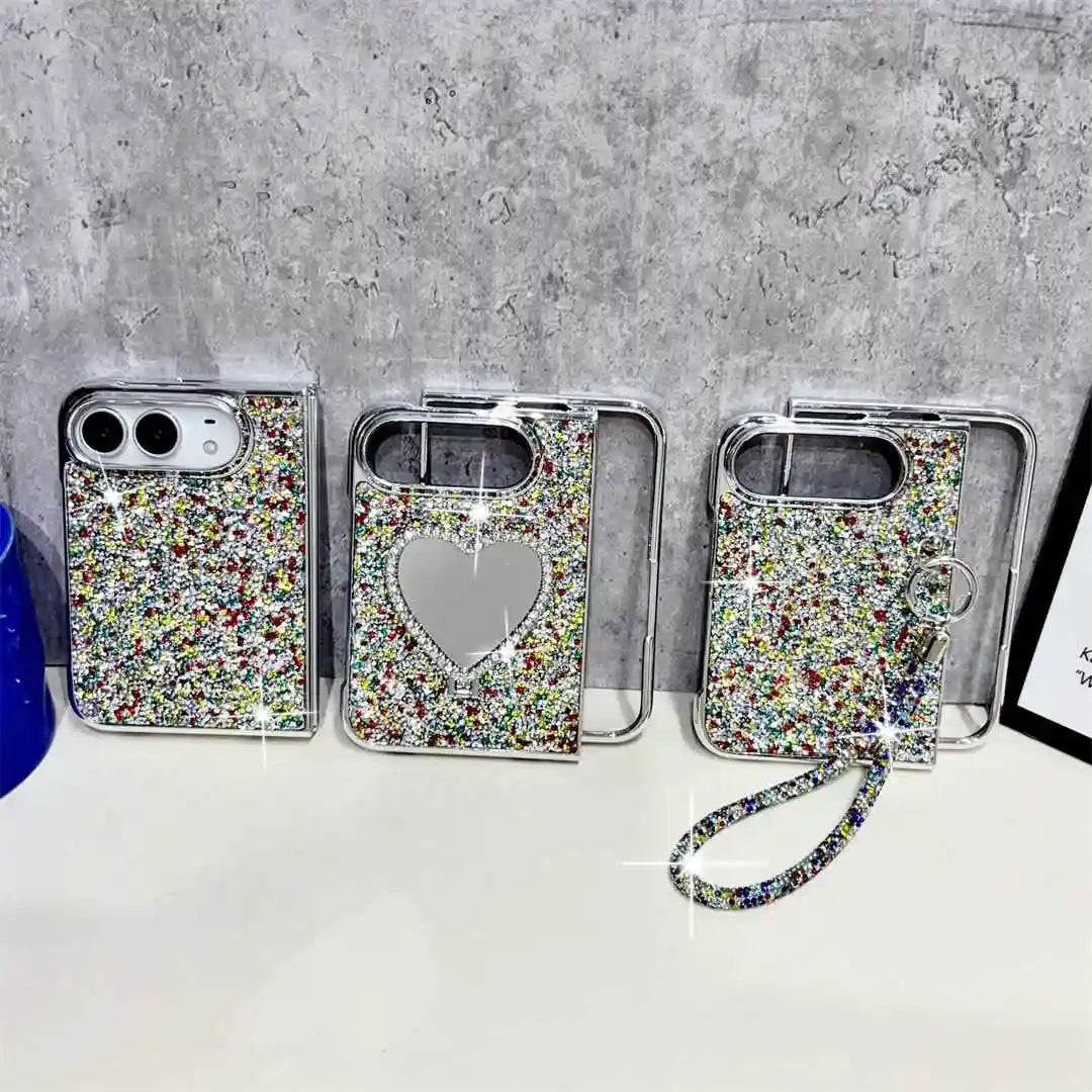 Three silver phone cases covered in multicolor rhinestones are displayed on a white surface against a gray concrete wall. One case features a heart-shaped mirror, and another has a matching rhinestone wrist strap.