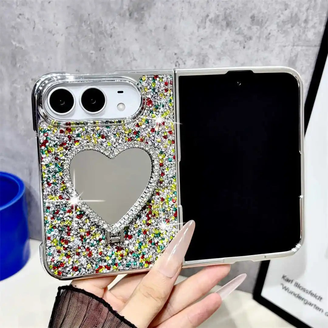 A hand holds a silver smartphone case decorated with multicolor glitter and a heart-shaped mirror on the back. The phone inside the case is black, and the background shows a gray textured wall and a blue cup.