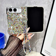 A hand holds a silver flip phone case covered in multicolor rhinestones and a matching wrist strap, shown next to an open black smartphone against a gray textured wall.