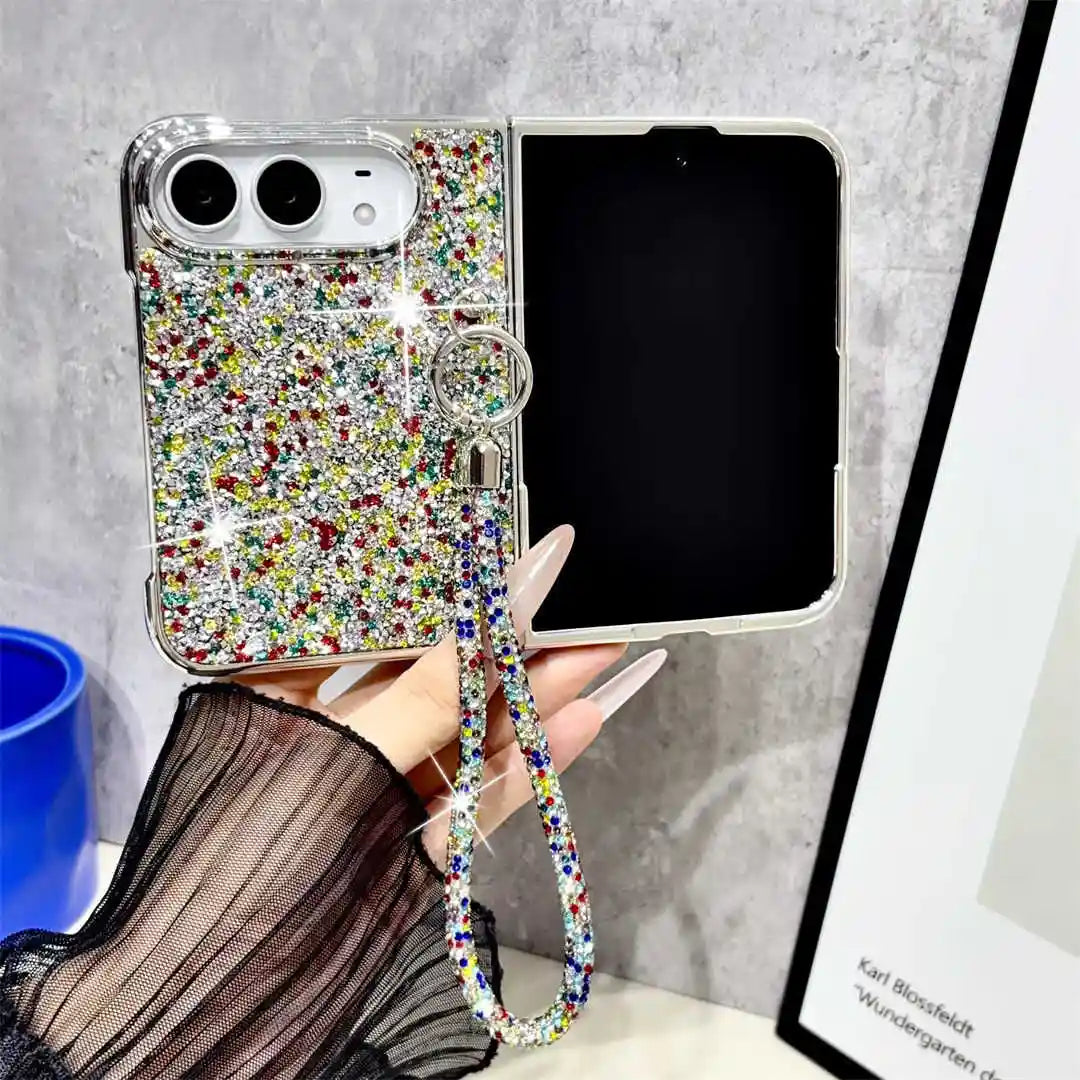 A hand holds a silver flip phone case covered in multicolor rhinestones and a matching wrist strap, shown next to an open black smartphone against a gray textured wall.