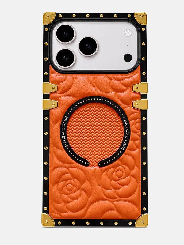 An orange embossed leather phone case with a rose pattern, gold-tone corner protectors, and a black studded border. It features a circular MagSafe-compatible ring on the back and a silver camera cutout at the top.