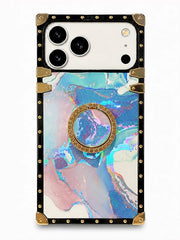 A protective phone case with a black border, gold-tone rivets and corner guards, featuring a holographic marble pattern in shades of blue, pink, and purple, with a built-in gold circular ring stand in the center.